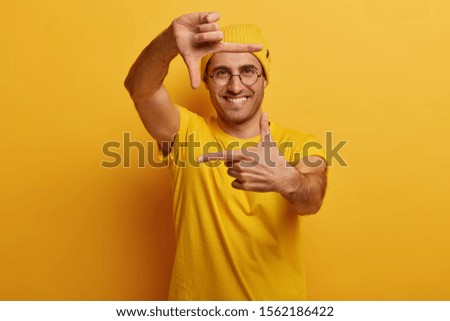 Fascinated young man with cheerful expression, makes hand frame, tries to find right perspective, pretends shooting scene, searches inspiration or right angle, wears yellow outfit, feels lucky