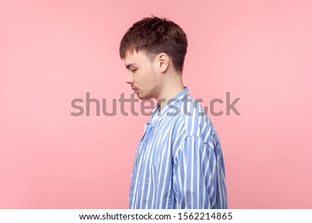 Side view portrait of upset brown-haired man with small beard and mustache in casual striped shirt standing looking down with bowed head, depression. indoor studio shot isolated on pink background