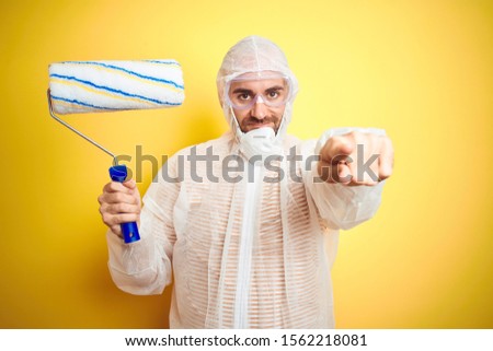 Young man wearing painter equipment and holding painting roller over isolated yellow background pointing with finger to the camera and to you, hand sign, positive and confident gesture from the front