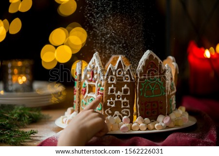Little gingerbread houses with glaze standing on table with red tablecloth and decorations, candles and lanterns. Lights of Christmas tree on background.  Powdered sugar falling like snow flakes