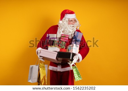 Male actor in a costume of Santa Claus holds a lot of gift boxes and packages in his hands and poses on a yellow background