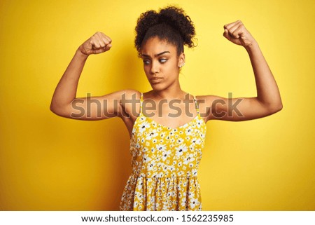 African american woman wearing casual floral dress standing over isolated yellow background showing arms muscles smiling proud. Fitness concept.