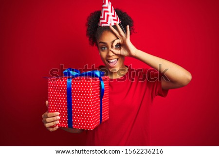 African american woman wearing funny hat holding birthday gift over isolated red background with happy face smiling doing ok sign with hand on eye looking through fingers