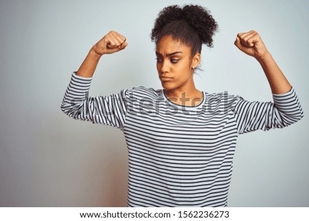 African american woman wearing navy striped t-shirt standing over isolated white background showing arms muscles smiling proud. Fitness concept.