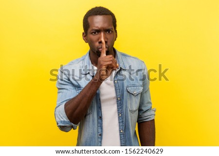 Shh, keep silence. Portrait of serious handsome man in denim casual shirt with rolled up sleeves gesturing to be quiet, asking for secrecy conspiracy. indoor studio shot isolated on yellow background