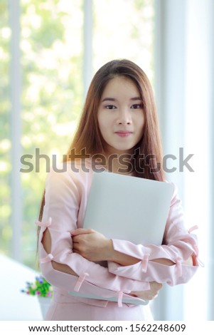 A beautiful young businesswoman, long hair in a pink dress on hand, has a laptop standing in the office with a smiling face ready to work.