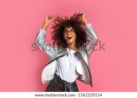 Happy ecstatic mix race woman dancing and playing with curly hairs on pink background. Wearing stylish winter silver jacket. 