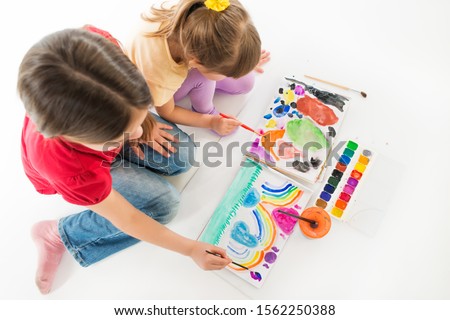 Two children in childrens clothes are sitting on the floor on their knees and enthusiastically painted with bright watercolor paints