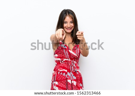 Caucasian girl in red dress over isolated white wall points finger at you while smiling