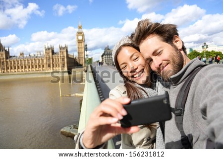 London tourist couple taking photo near Big Ben. Sightseeing woman and man having fun using smartphone camera smiling happy near Palace of Westminster, Westminster Bridge, London, England.