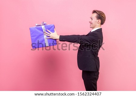 Take this gift! Side view of happy generous man in suit and with stylish haircut holding out gift box and smiling, giving present with joy, charity concept. studio shot isolated on pink background