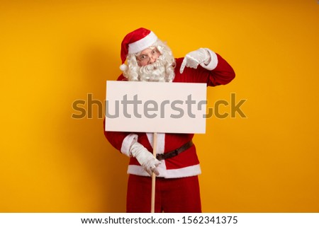 Emotional male actor in a costume of Santa Claus holds in his hands white billboard on a stick for recording ads and posing on a yellow background