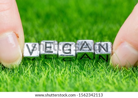 Close up on word VEGAN written in metal letters laid on grass and held between the fingers of a woman. Concept of alimentation background