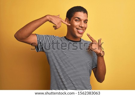 Young handsome arab man wearing navy striped t-shirt over isolated yellow background smiling cheerful showing and pointing with fingers teeth and mouth. Dental health concept.