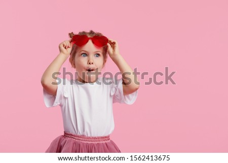 Portrait of surprised cute little toddler girl in the heart shape sunglasses. Child with open mouth having fun isolated over pink background. Looking at camera. Wow funny face Royalty-Free Stock Photo #1562413675