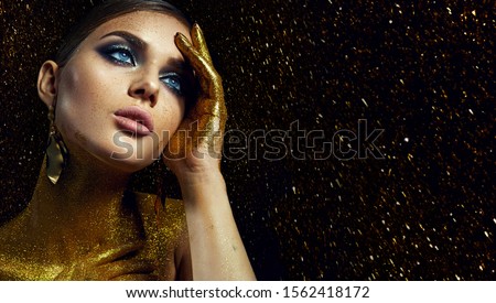 Face and hands of a beautiful showy young girl with bright makeup and combed hair in shiny gold sparkles on a black background.