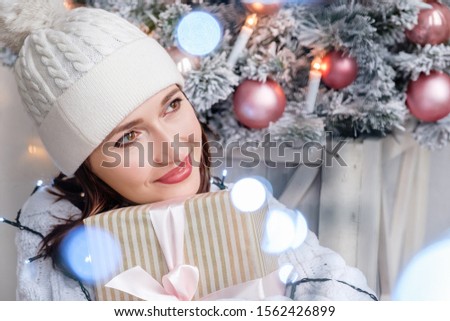 Beautiful positive dreamy woman in white sweater and knitted hat hugs her gift while sitting on background of Christmas tree among the LED lights. Concept of New Year gifts for promotions and sales