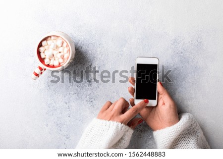 Female hands hold a cup of coffee with marshmallows and mobile smart phone .Winter Holiday concept. Winter Warm Drink Hot chocolate.Modern technology Flat lay, top view.