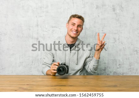 Young photographer holding a camera on a table joyful and carefree showing a peace symbol with fingers.