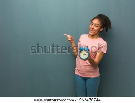 Young black woman pointing to the side with finger. She is holding an alarm clock.