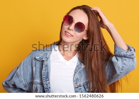Horizontal indoor shot of cheerful dreaming young girl posing isolated over yellow background in studio, putting one hand on head, having long hair, wearing accessories. People and emotions concept.