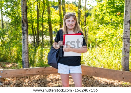 Happy/smiling teen girl standing in a park near her school during sunset/golden hour while holding binders and wearing a backpack.