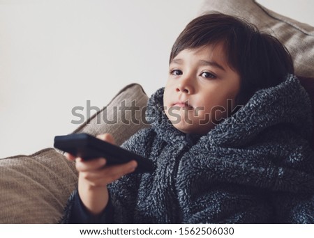 Cute kid sitting on sofa holding remote control, A happy child boy relaxing at home watching TV during cold weather outside in Autumn or Winter. Indoors activities for kids during bad weather 