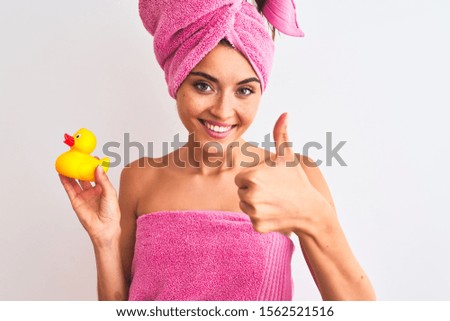 Young beautiful woman wearing shower towel holding duck over isolated white background happy with big smile doing ok sign, thumb up with fingers, excellent sign