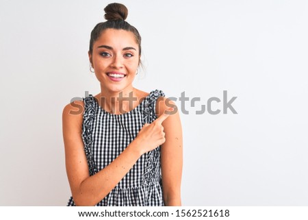 Beautiful woman with bun wearing casual dresss standing over isolated white background cheerful with a smile on face pointing with hand and finger up to the side with happy and natural expression