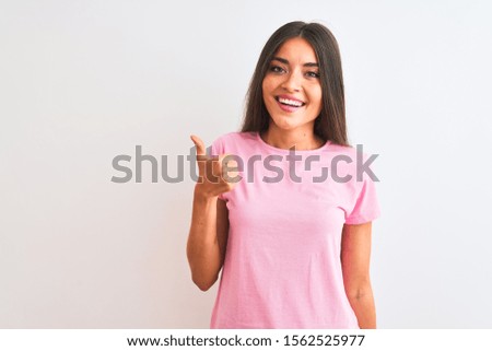 Young beautiful woman wearing pink casual t-shirt standing over isolated white background doing happy thumbs up gesture with hand. Approving expression looking at the camera showing success.