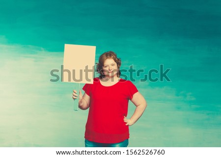 Young attractive plump woman with red hair holds a blank poster on a blue background.