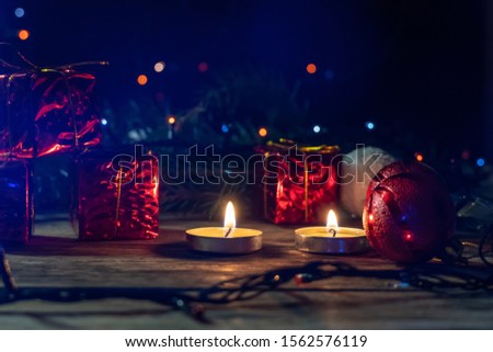 Red mini gifts on a wooden background among fir branches, lighted candles in a dark room. Beautiful, magical Christmas background with copy space.