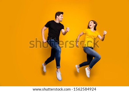 Full size photo of sporty guy and lady couple jumping high active way of life pair marathon participants wear casual jeans black t-shirts isolated yellow color background
