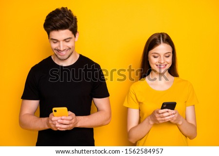 Photo of cheerful positive cute nice addicted people browsing through their telephones chatting with each other despite standing side by side isolated vivid shiny color background in black t-shirt