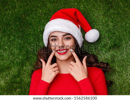 From above optimistic female in Santa hat making smile with index fingers while lying on green grass during Christmas celebration