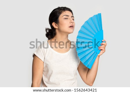 Tired exhausted Indian girl waving blue paper fan standing isolated on grey background, suffering from heat, sweaty young woman cooling in hot summer weather, high temperature, close up Royalty-Free Stock Photo #1562643421