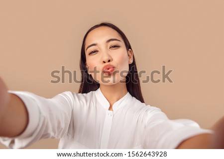 Young woman wearing white shirt with dark long hair standing isolated on bage background taking selfie photo on smartphone pouting lips kissing camera close-up