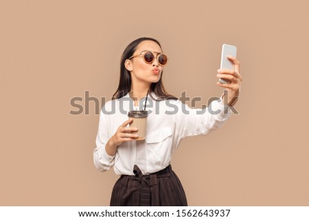 Young woman wearing white shirt and sunglasses with dark long hair standing isolated on bage background holding cup of hot coffee holding smartphone taking selfie photo posing to camera pouting lips