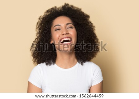 Head shot excited African American girl laughing out loud at funny joke, feeling positive, having fun alone, happy beautiful young female in white t-shirt standing isolated on brown background