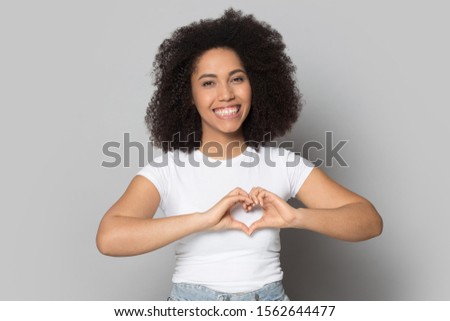 Head shot smiling African American young female with toothy smile showing heart gesture with fingers, happy girl wearing white t-shirt looking at camera, standing isolated on grey background