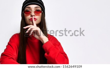 Hipster girl in a fashionable black hat headdress with long hair, showing hush, shhh, secrecy, silent sign, isolated on light studio wall background