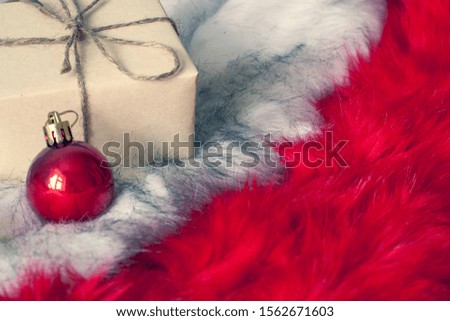 Gift with a bow in craft gray paper on fluffy white and red fur. A small red Christmas ball is nearby. Selective focus on the gift. Copy space.