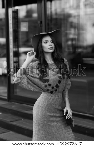 Attractive long-haired woman wearing stylish gray maxi dress, wide-brimmed hat, high-heeled boots, holding handbag. Young female model in trendy outfit posing on street outside exquisite building.