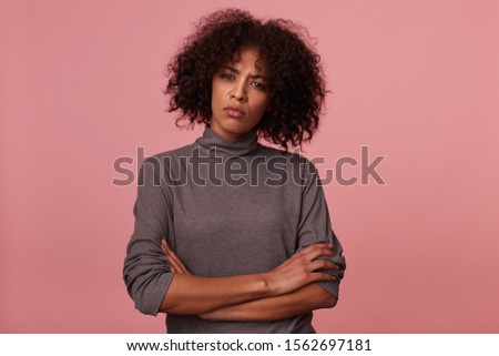 Severe young dark skinned brunette female with short haircut folding hands on her chest while standing against pink background, looking seriously to camera and frowning her eyebrows