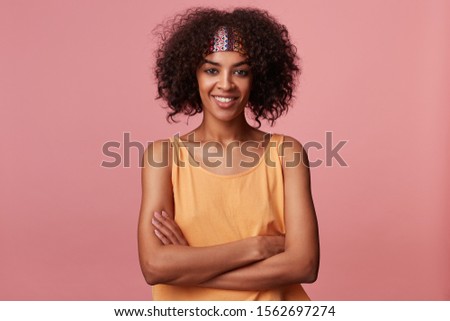 Portrait of cheerful attractive young dark skinned lady with short curly brown hair looking positively at camera with pleasant smile, folding hands on her chest while standing over pink background