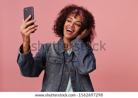 Joyful lovely smiling dark skinned curly brunette lady with short haircut wearing colorful headband while posing over pink background, raising hand with mobile phone while making photo of herself