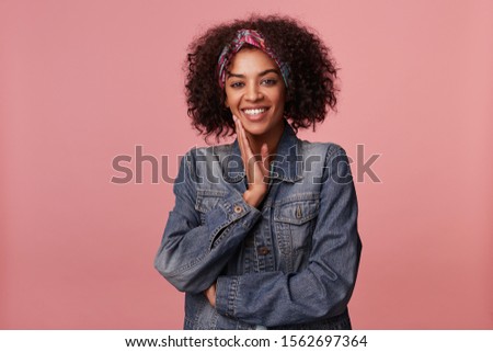 Joyful attractive young dark skinned lady with short curly brown hair wearing colorful headband and touching gently her face with raised palm, smiling widely to camera over pink background