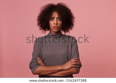 Happy attractive young dark skinned brunette woman with short curly hair posing over pink background in grey throat sweater, smiling widely to camera and holding raised hands on her head