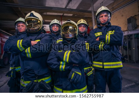 Portrait of group firefighters standing proudly in front of firetruck inside the fire station wearing full protective gear with helmets on