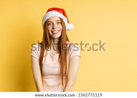 Excited ginger young caucasian lady in casual clothes and Santa hat having happy look, celebrating victory, rejoicing christmas holidays clenching fists jumping up and down over yellow background.